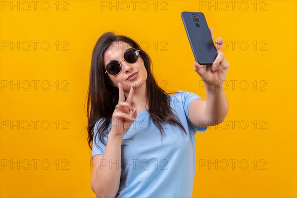 Studio portrait with yellow background of a casual woman with sunglasses taking a selfie with mobile phone