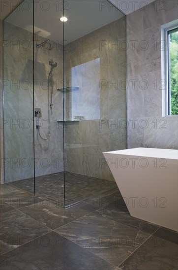Clear glass shower stall and white freestanding vessel shaped bathtub in bathroom with grey ceramic tile floor and walls on ground floor inside modern cubist style home, Quebec, Canada, North America