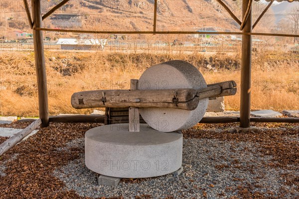 Old antique millstone and wheel in covered shelter with mountain community in background in South Korea