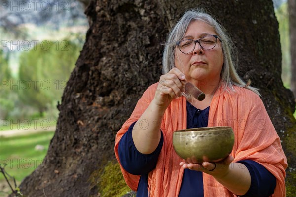 Mature woman with white hair meditating in the forest with her eyes closed and a Tibetan singing bowl ringing