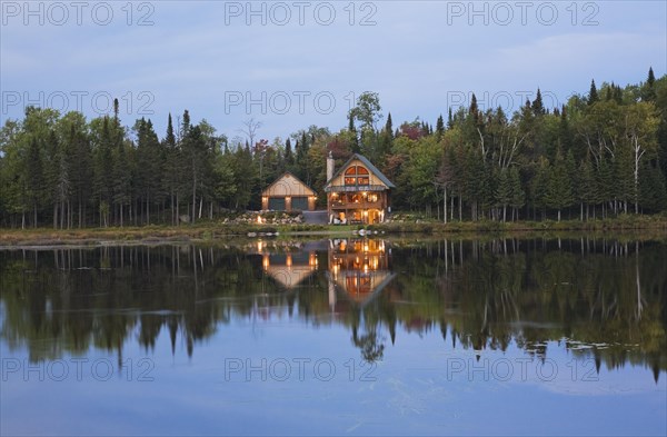 Illuminated two car garage and handcrafted two story spruce log cabin home with fieldstone chimney and green sheet metal roof on edge of lake at dusk in autumn, Quebec, Canada, North America