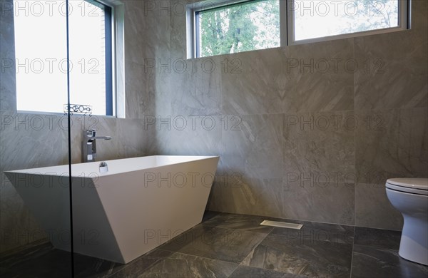 White freestanding vessel shaped bathtub in bathroom with grey ceramic tile floor and walls on ground floor inside modern cubist style home, Quebec, Canada, North America