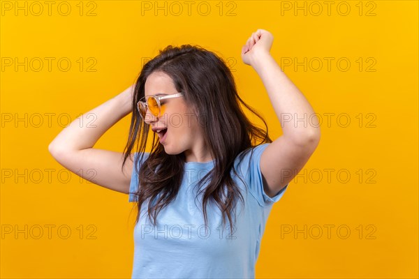 Studio portrait with yellow background of a happy woman wearing sunglasses dancing and celebrating raising arms
