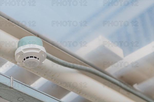 Electronic smoke detector on ceiling of enclosed pedestrian walkway with blurred plexiglass window in background in South Korea
