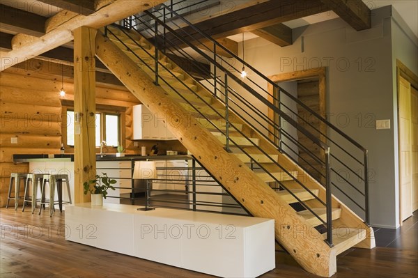 Kitchen with white wooden cabinets, metal bar stools and wooden log stairs with black wrought iron railings and long cabinet table inside contemporary style log home, Quebec, Canada, North America