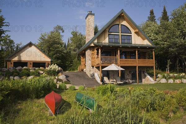 Garage and handcrafted two story spruce log home cabin with fieldstone chimney, green sheet metal roof and red and green canoes in summer, Quebec, Canada, North America