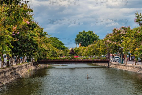 A tranquil water canal with a bridge, flanked by trees and clouds reflecting on the surface, in Chiang Mai, Thailand, Asia