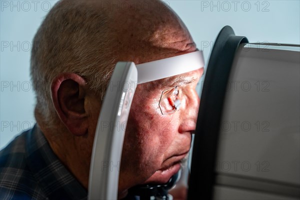 Close-up photo with side view of a senior man with eye opener leaning on front of a innovative machine during a laser treatment for glaucoma
