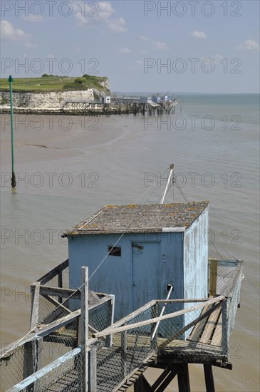 A tranquil beach scene with a blue hut on a wooden pier, under a sunny sky with a calm sea and coastline in the backgroundFishery in Talmon. editorial