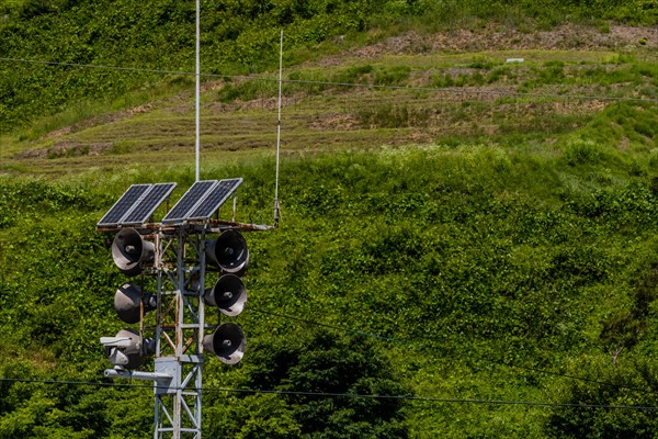 Communication tower consisting of loud speakers, solar panel and closed circuit camera with tree covered mountain in background in South Korea