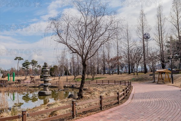 A peaceful park scene with a pond, leafless trees and a brick footpath under a cloudy sky, in South Korea