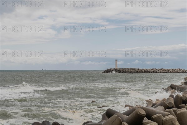 Waves crash against the breakwater with a lighthouse in the distance under a gray sky, in Ulsan, South Korea, Asia
