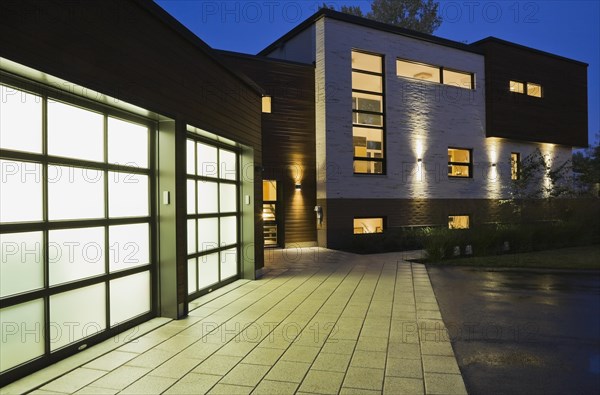 Illuminated two car garage and beige stone with brown cedar wood siding modern cubist style home facade with paving stone and black asphalt driveway at dusk in summer, Quebec, Canada, North America