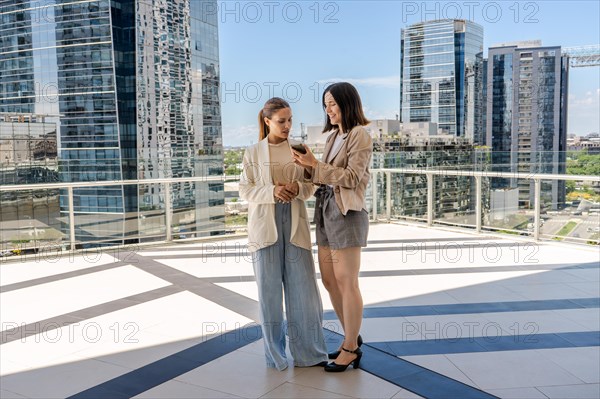 Two businesswomen standing on a terrace, one of them holding a cell phone and showing an email to her colleague. The scene is set in a city with tall buildings in the background