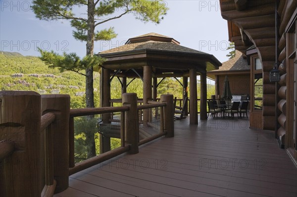 Brown stained gazebo on wooden deck with tempered glass and log railings attached to back of luxurious log cabin home overlooking Mt-Tremblant ski resort in summer, Quebec, Canada, North America
