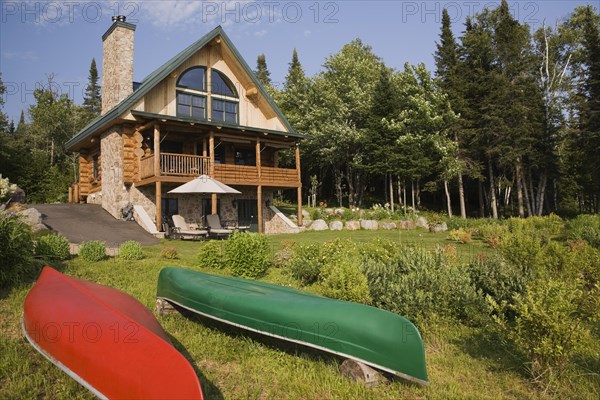Handcrafted two story spruce log home cabin with fieldstone chimney, green sheet metal roof and red and green canoes in summer, Lantier, Lanaudiere, Quebec, Canada, North America