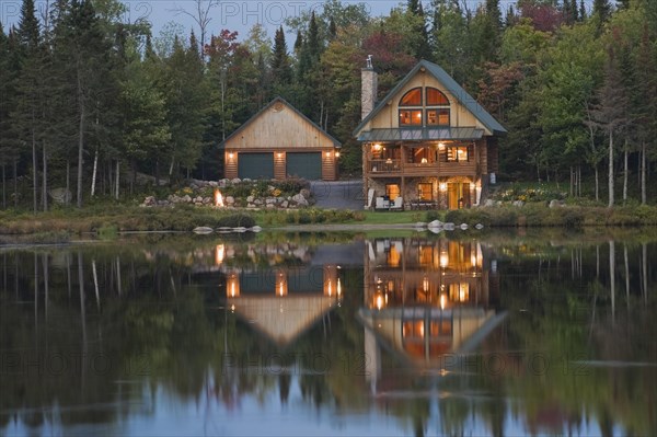 Illuminated two car garage and handcrafted two story spruce log cabin home with fieldstone chimney and green sheet metal roof on edge of lake at dusk in autumn, Quebec, Canada, North America
