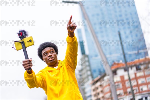 African young vlogger pointing ahead during an online video with a mobile attached on mic and tripod in the city