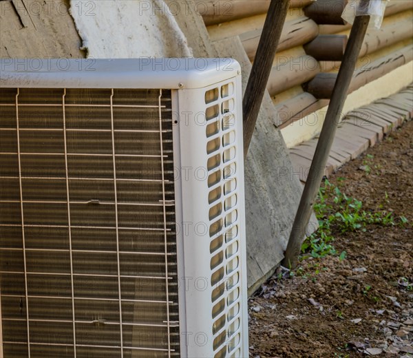Closeup of air conditioner condenser unit on ground with exterior wall of log cabin in background in South Korea
