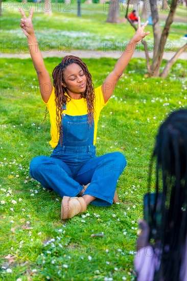 Vertical photo of an African young woman wearing dungarees sitting with arms raised while friend taking a photo in the park