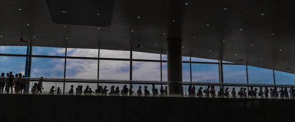 Silhouette of people standing in line on upper floor of airport building in front of blue sky outside window in China