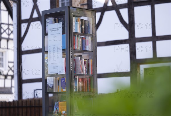 Open bookcase in front of half-timbered house and fence, many colourful books in a glass display case, outdoors, in the foreground on the right, a green plant has disappeared, Volmarstein, Ruhr area, Germany, Europe