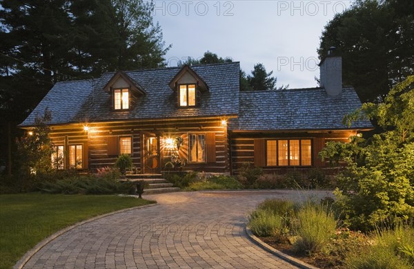Illuminated old 1800s reconstructed log cabin home facade and paving stone driveway at dusk, Quebec, Canada, North America