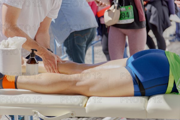 An athletic trainer wearing a white t-shirt is providing a professional leg massage to a runner reclining on a massage table, focusing on the calf muscles, during a marathon event. The runner, dressed in athletic clothing, appears to be receiving post-race care outdoors with other participants and spectators in the background