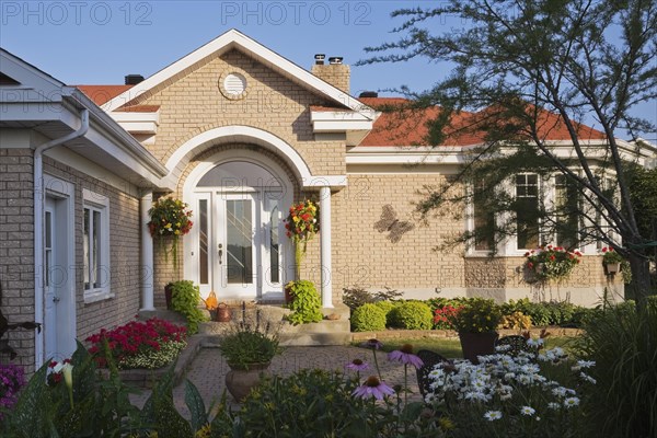 Tan brick home with white trim and landscaped front yard with raised border with red Pelargonium, Geranium flowers, Echinacea purpurea, Coneflowers and white Leucanthemum, Oxeye daisies in summer, Quebec, Canada, North America