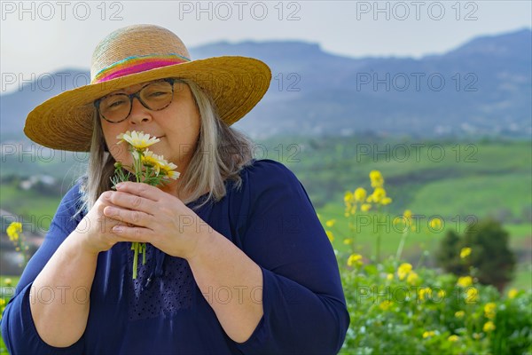 Mature woman with white hair and hat holding a bouquet of daisies in her hands in the background a mountain landscape
