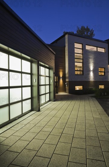 Illuminated two car garage and beige stone with brown cedar wood siding modern cubist style home facade with paving stone and black asphalt driveway at dusk in summer, Quebec, Canada, North America