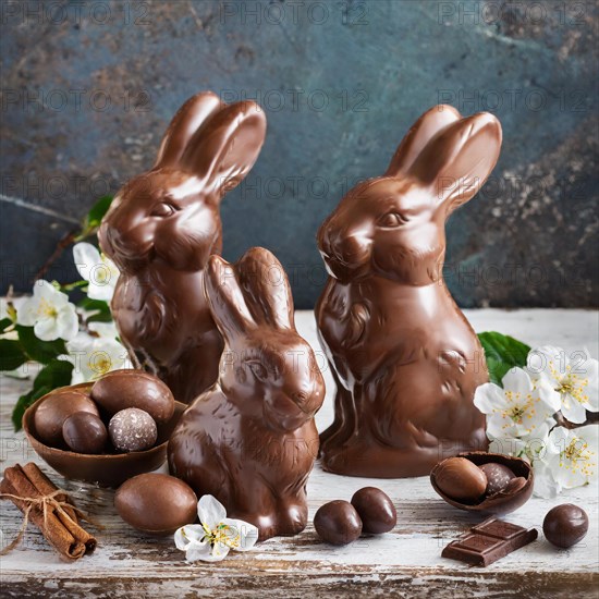 Three chocolate bunnies next to chocolate eggs and cinnamon sticks, surrounded by fresh flowers, symbolising Easter, AI generated, AI generated