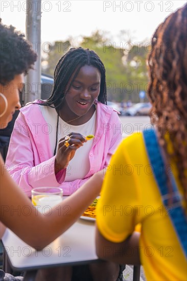 Vertical photo of three young african female friends eating nachos in a sidewalk restaurant