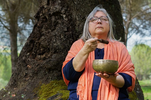 Mature gray-haired woman sitting in a tree in the forest with her eyes closed, ringing a Tibetan singing bowl