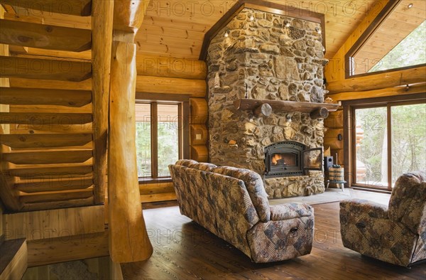 Wooden log stairs and upholstered sofas with lit fieldstone and porous rock fireplace in living room inside handcrafted red cedar log cabin home, Quebec, Canada, North America