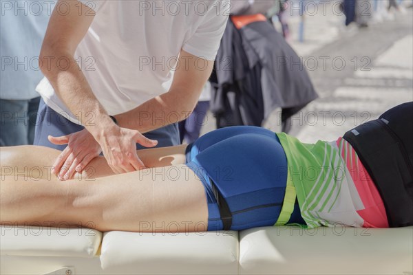 A first responder is administering first aid to a marathon runner who has collapsed on the ground. The rescuer is pressing with both hands on the runners back, possibly performing a massage or assessing the injury. The runner, dressed in athletic gear, lies prone on a white mat, suggesting a quick response to an incident during the event