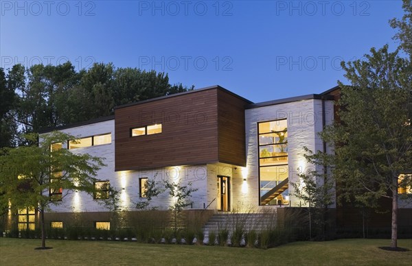 Illuminated beige stone with brown cedar wood siding modern cubist style home with landscaped front yard at dusk in summer, Quebec, Canada, North America