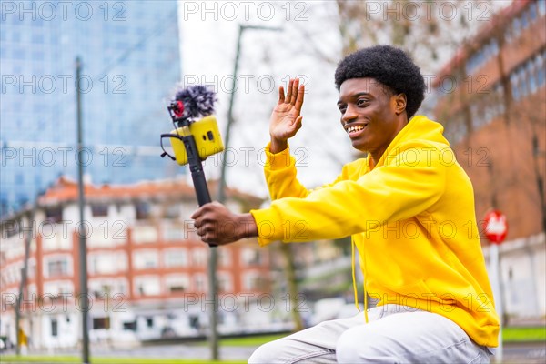 Side view photo of a young african stylish vlogger recording video and waving in the city