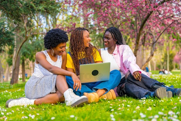 Happy african female friends using laptop sitting on a park in spring