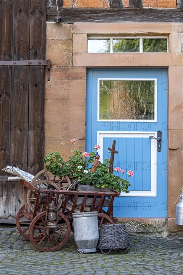 Curious decoration on a front door, flower pot, geraniums, old wagon with firewood, milk can and basket, idyll, old town, Ortenberg, Wetterau, Vogelsberg, Hesse, Germany, Europe