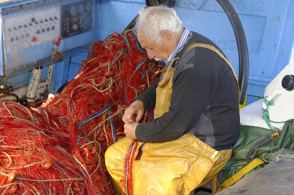 Focussed fisherman in yellow working clothes repairs red nets on board a boat, Marseille, Departement Bouches-du-Rhone, Provence-Alpes-Cote d'Azur region, France, Europe
