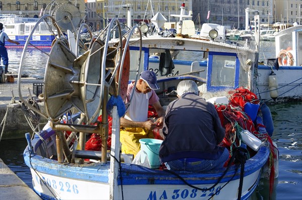 A fisherman repairs nets on his blue boat in the quiet harbour during daylight, Marseille, Departement Bouches-du-Rhone, Provence-Alpes-Cote d'Azur region, France, Europe
