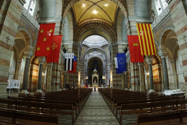 Marseille Cathedral or Cathedrale Sainte-Marie-Majeure de Marseille, 1852-1896, Marseille, View through the nave of a church with pews and various flags on the pillars, Interior view of a church with arches, flags and a row of pews, Marseille, Departement Bouches du Rhone, Region Provence Alpes Cote d'Azur, France, Europe