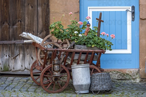 Curious decoration on a front door, flower pot, geraniums, old wagon with firewood, milk can and basket, idyll, old town, Ortenberg, Wetterau, Vogelsberg, Hesse, Germany, Europe