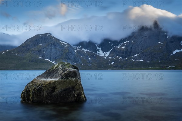 Landscape with sea and mountains on the Lofoten, view over the fjord Flakstadpollen to the mountains Stortinden and others. A rock in the foreground. At night at the time of the midnight sun, clouds move over the mountain peaks. Long exposure. Early summer. Flakstadoya, Lofoten, Norway, Europe