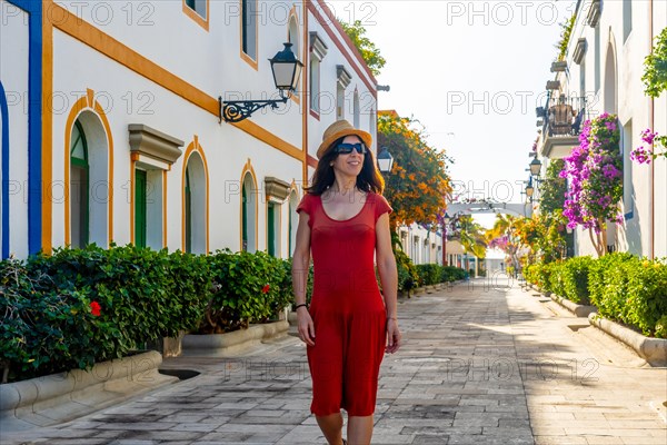 A woman in a red dress is walking down a street with a hat on. The street is lined with buildings and has a lot of greenery. The woman is enjoying her walk and taking in the scenery