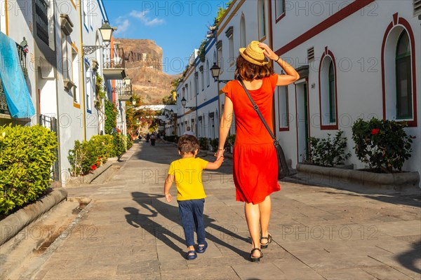 A woman and a child walk down a narrow street in a foreign city. The woman is wearing a red dress and a straw hat. The scene is lively and colorful, with many people walking around
