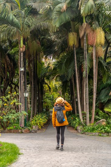 A woman wearing a yellow jacket and a straw hat walks through a forest. She is carrying a backpack and a cell phone