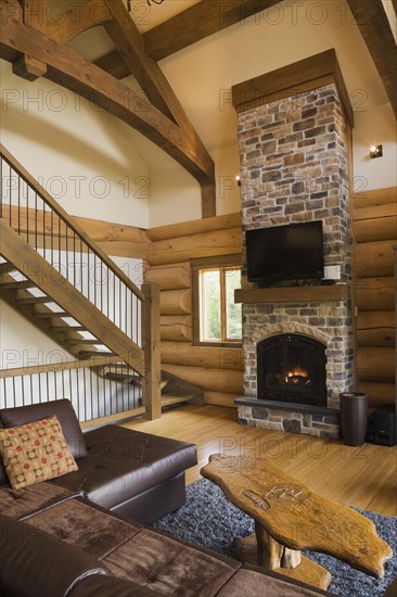 Brown leather L-shaped sofa and wooden coffee table in front of lit nuanced tan and brown natural stone propane gas fireplace next to wooden stairs with wrought iron handrail leading to upstairs floor in living room with hardwood floor inside luxurious contemporary Scandinavian style log cabin home, Quebec, Canada, North America
