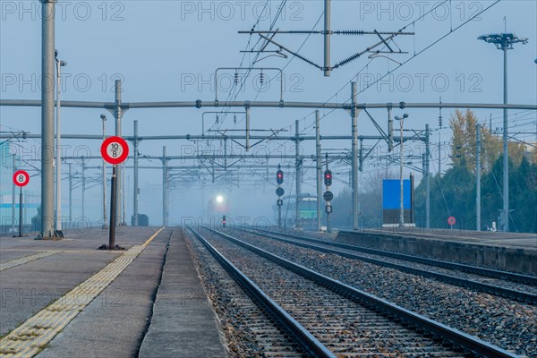 Empty train platform at a station on a foggy day with overhead electrical equipment and light of approaching train in distance in South Korea